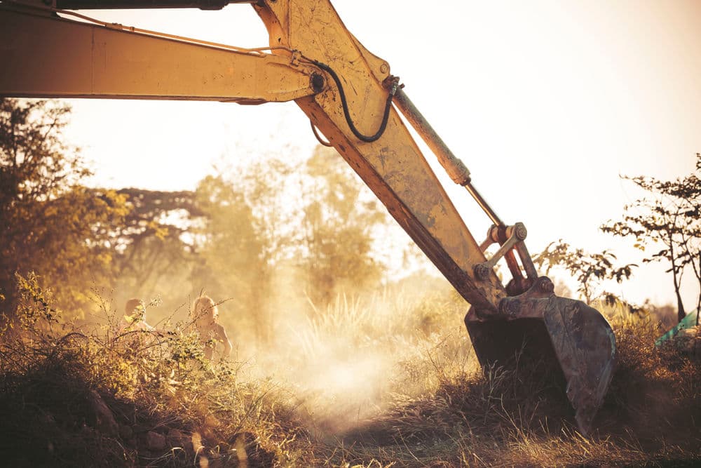 Excavator working at sunset with dust and two people in the background.